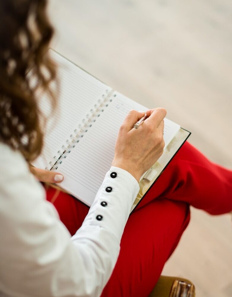A woman in a white shirt and red trousers writing in a notebook